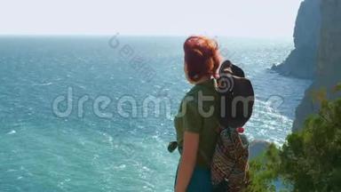 年轻的红发女孩旅行者戴着牛仔帽，背着背包站在山顶上，看着蓝色的大海。